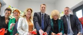 W białym korytarzu stoi minister Czarnek, dwóch mężczyzn w garniturach i trzy kobiety - ubrane na czarno, zielono i biało, wszyscy trzymają w rękach nożyczki i przecinają czerwoną wstęgę.