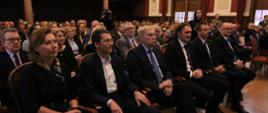 Zdjęcie z boku na salę, na ustawionych w rzędy krzesłach siedzą elegancko ubrani ludzie, w tle czerwono-beżowa ściana i dwa okna.