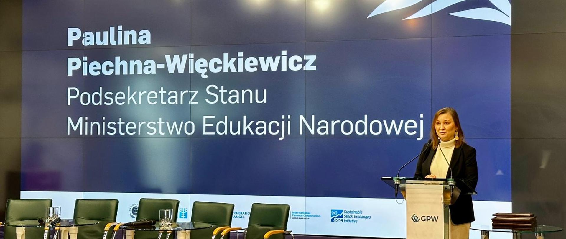 Wiceminister Paulina Piechna-Więckiewicz stoi przy mównicy i mówi do mikrofonu w tle duży ekran z napisem Paulina Piechna-Więckiewicz Podsekretarz Stanu Ministerstwo Edukacji Narodowej