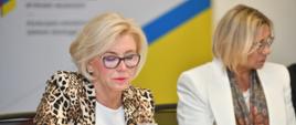 Wiceminister Machałek w marynarce w brązowo-żółto-biały drobny wzorek siedzi za białym stołem i pisze na kartce, obok niej kobieta w białej marynarce.