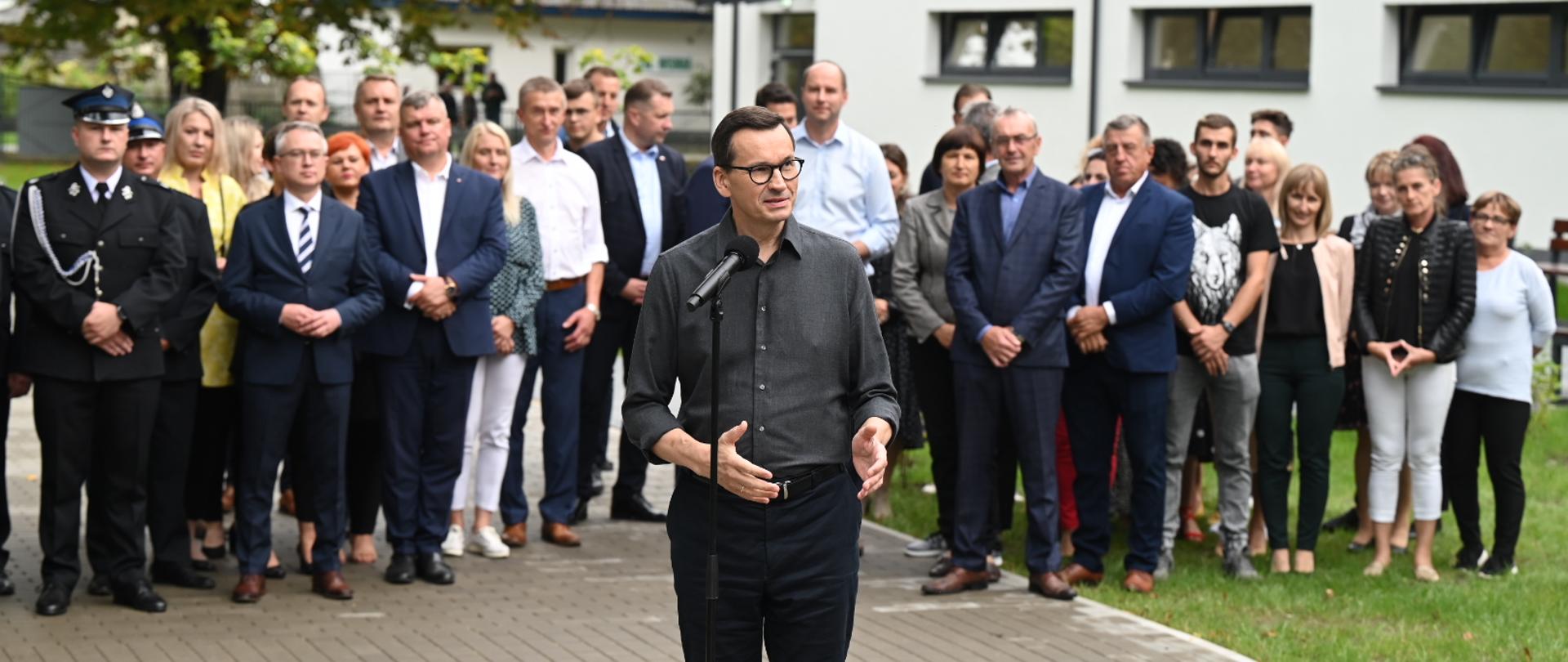 Mężczyzna w szarej koszuli i okularach stoi na wyłożonym szarą kostką chodniku i mówi do mikrofonu na stojaku, za nim duża grupa ludzi, za nimi z prawej biały budynek.