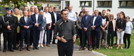 Mężczyzna w szarej koszuli i okularach stoi na wyłożonym szarą kostką chodniku i mówi do mikrofonu na stojaku, za nim duża grupa ludzi, za nimi z prawej biały budynek.