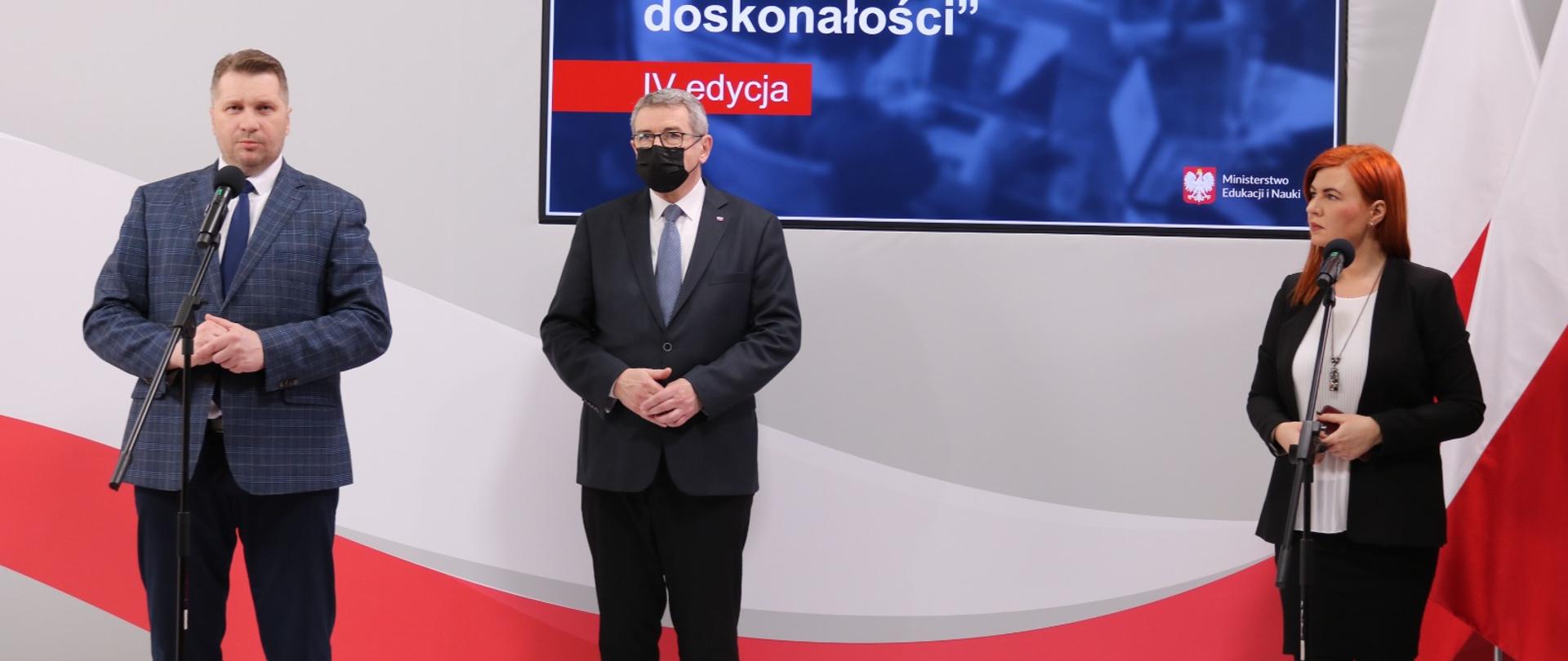 Minister Przemysław Czarnek, wiceminister Wojciech Murdzek oraz rzecznik prasowa MEiN Anna Ostrowska stoją podczas konferencji, za nimi znajduje się telebim.