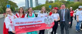 Widok na ulicę, kilka dziewczyn w kolorowych strojach niesie rozpostartą polska flagę z napisem Polish Student Alliance, obok nich wiceminister Rzymkowski, w tle wysokie budynki.