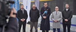 Na zdjęciu wiceminister Jan Szyszko w otoczeniu innych uczestników spotkania przed mikrofonami odpowiada na pytania dziennikarzy. W tle widać wagon pociągu osobowego.