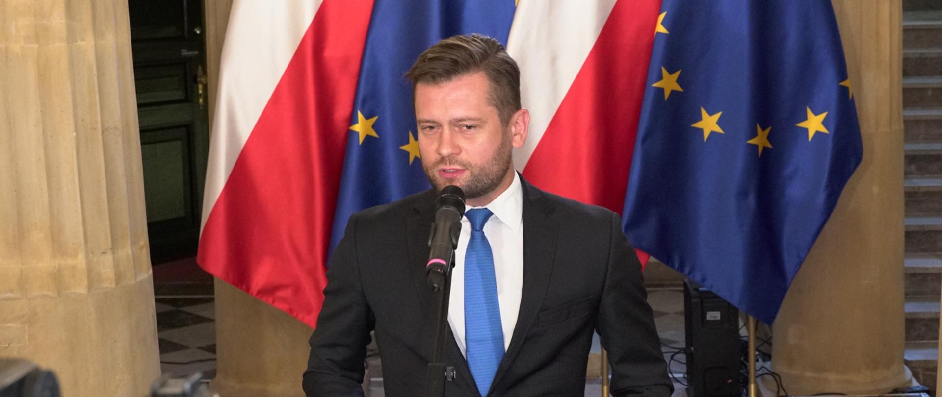 Na zdjęciu znajduje się ubrany w ciemny garnituru, białą koszulę i błękitny krawat mężczyzna. Jest on w trakcie wystąpienia publicznego. W tle znajdują się cztery flagi, ustawione naprzemiennie: biało-czerwone oraz Unii Europejskiej (granatowe z żółtymi gwiazdami).