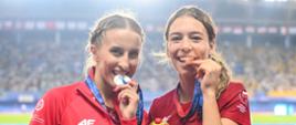 Dwie kobiety w czerwonych sportowych bluzach trzymają małe maskotki - misie panda, i gryzą medale na wstążkach.