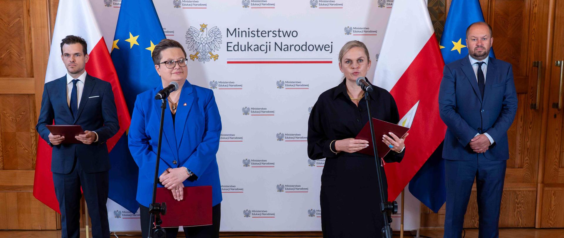 Na tle polskiej i unijnej flagi, na jasnym tle z logami Ministerstwa Edukacji Narodowej, stoi czworo dorosłych. Po lewej stronie od środka, kobieta w okularach, niebieskiej marynarce i czarnych spodniach stoi z mikrofonem przy ustach, trzymając czerwoną teczkę w lewej ręce. Obok niej stoi mężczyzna w granatowym garniturze z krawatem, trzymający czerwoną teczkę.
Po prawej stronie od środka, kobieta z blond włosami, ubrana w czarną sukienkę, trzyma mikrofon i czerwoną teczkę. Obok niej stoi mężczyzna z brodą i łysą głową, w niebieskim garniturze i krawacie. Oświetlenie jest jasne i równomierne. Całość wygląda jak oficjalna konferencja prasowa.