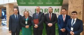 Na tle białego budynku stoi zielono-biała ścianka z napisem FORUM, przed nią pięciu mężczyzn w garniturach i kobieta w czarnej marynarce.