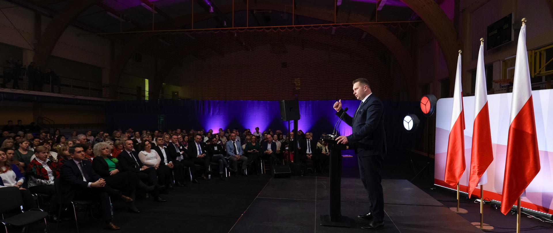 Widok z boku na dużą zaciemnioną wypełnioną ludźmi salę, przed nimi na oświetlonym podwyższeniu stoi przy mównicy minister Czarnek i mówi do mikrofonu, za nim rząd polskich flag.