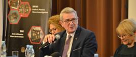 Za nakrytym niebieskim materiałem stołem siedzi uśmiechnięty wiceminister Murdzek i mówi do trzymanego w ręku mikrofonu, za nim żółta ściana częściowo przysłonięta brązową zasłoną, z obu stron siedzą dwie kobiety.