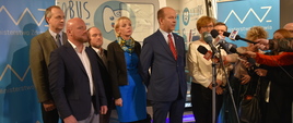 Konferencja prasowa. Minister zdrowia Konstanty Radziwiłł przemawia na tle jednego z dentobusów. Obok niego stoją wiceminister zdrowia Józefa Szczurek-Żelazko, a także przedstawicieli wykonawcy oraz posłowie Ziemi Radomskiej. 