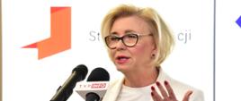 Wiceminister Machałek w białym ubraniu stoi i mówi do trzech mikrofonów na stojakach, za nią biała ściana.