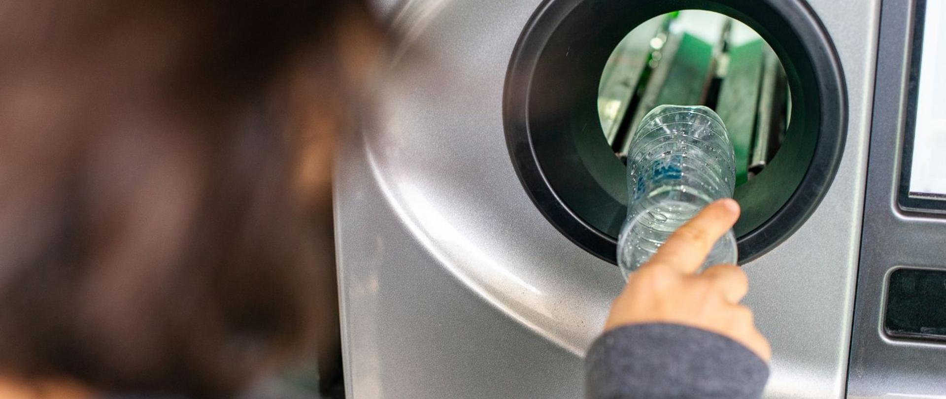 na zdjęciu widnieje postać kobiety, która wrzuca plastikową butelkę do automatu systemu kaucyjnego. 
