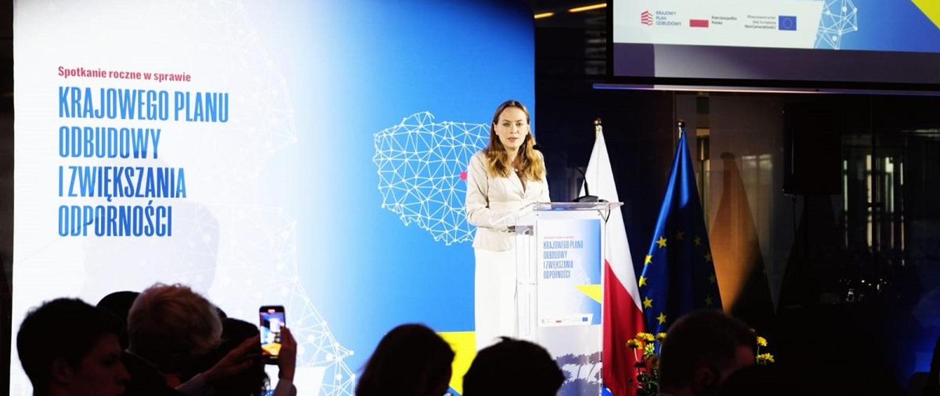 Minister funduszy i polityki regionalnej Katarzyna Pełczyńska-Nałęcz stoi przy mównicy i przemawia przez mikrofon, za jej plecami baner z napisem "Krajowy Plan Odbudowy i Zwiększania Odporności" oraz mapą Polski