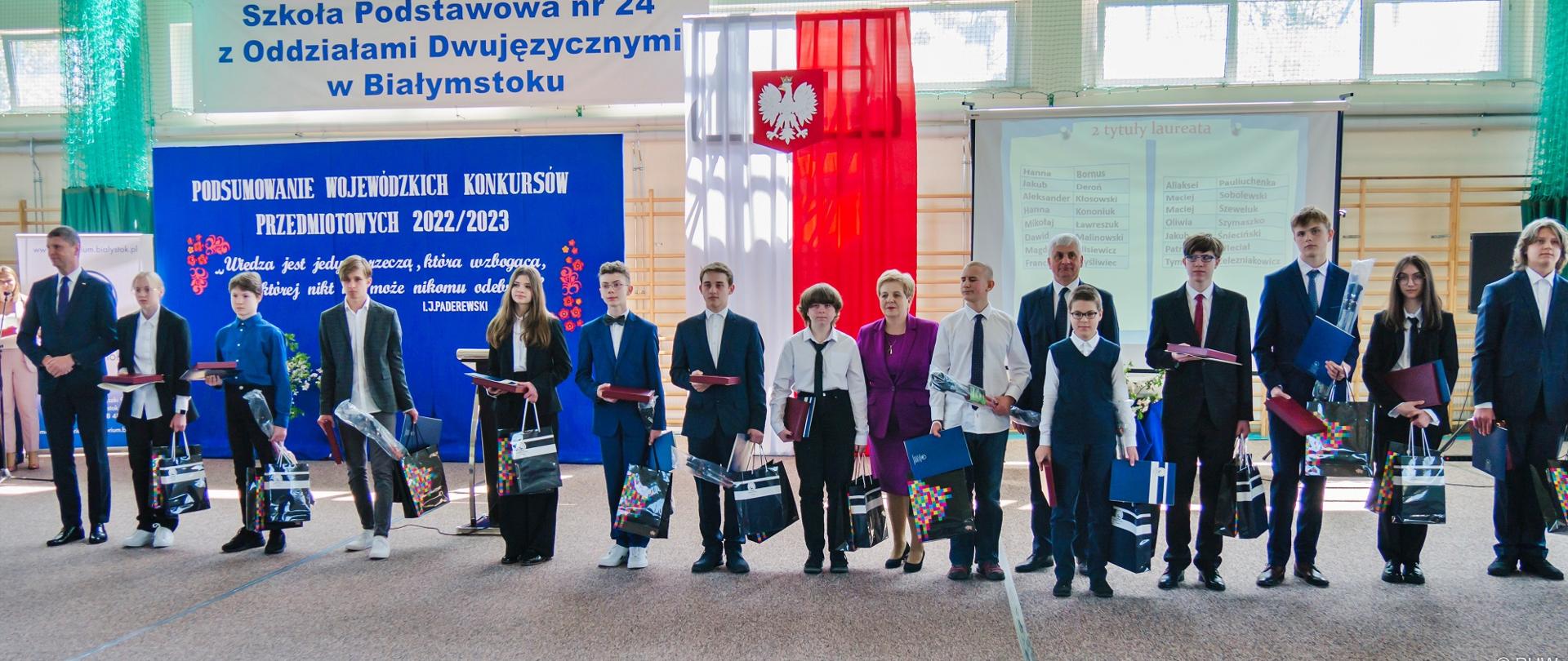 Na sali gimnastycznej stoi rząd elegancko ubranych osób, za nimi ściana z drewnianymi drabinkami przesłonięta granatowym materiałem z białym napisem Podsumowanie wojewódzkich konkursów przedmiotowych 2022/2023, nad nim napis Szkoła Podstawowa nr 24 z oddziałami dwujęzycznymi w Białymstoku, obok polska flaga.