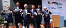 Na dworze w ostrym słońcu stoi grupa mężczyzn w garniturach, każdy trzyma mały kawałek wstęgi z napisem SOWA, przed nimi na ziemi siedzi grupa dzieci.