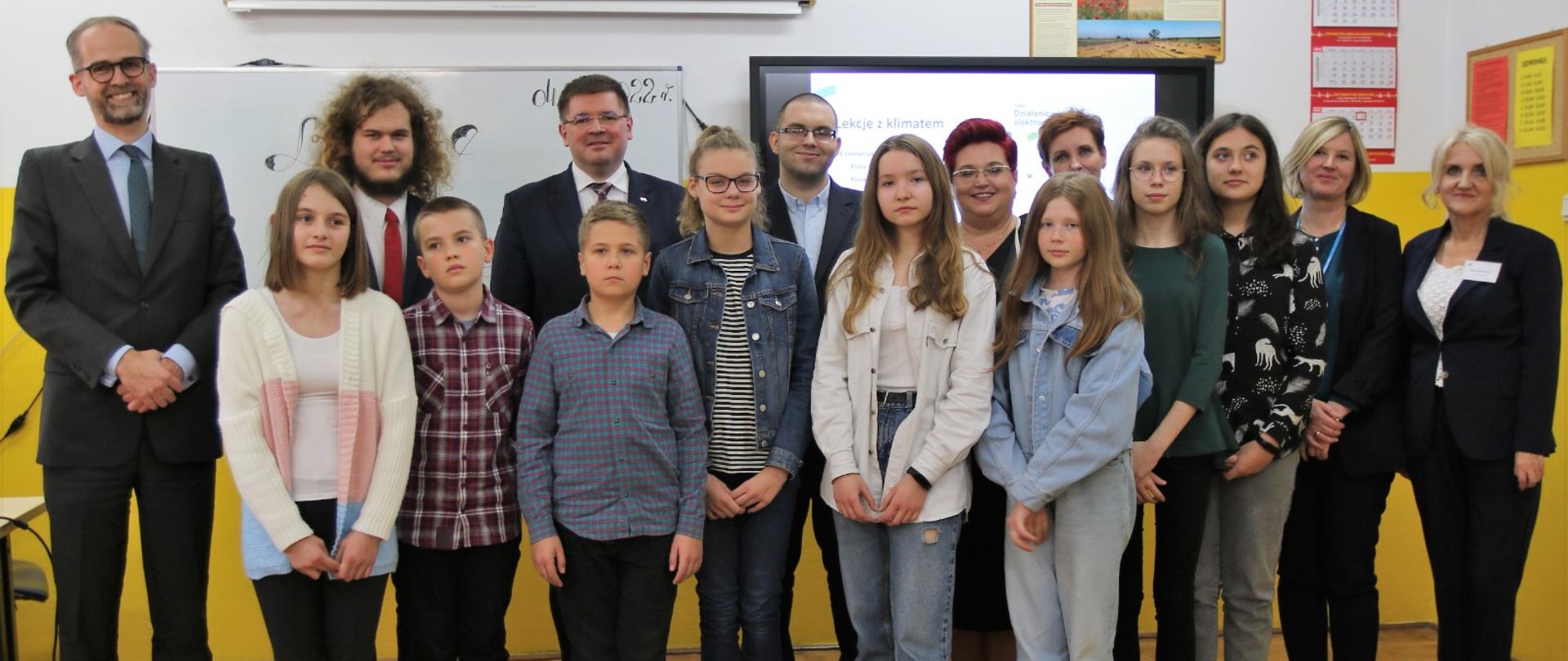Grupa dzieci i dorosłych stoi w rzędzie, za nimi duży monitor.