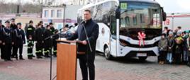 Na wyłożonym kostką podwórku stoi przy drewnianej mównicy minister Czarnek i mówi do kilku mikrofonów na stojakach, za nim biały autobus, grupa ludzi w czarnych mundurach, z drugiej strony grupa kolorowo ubranych dzieci.