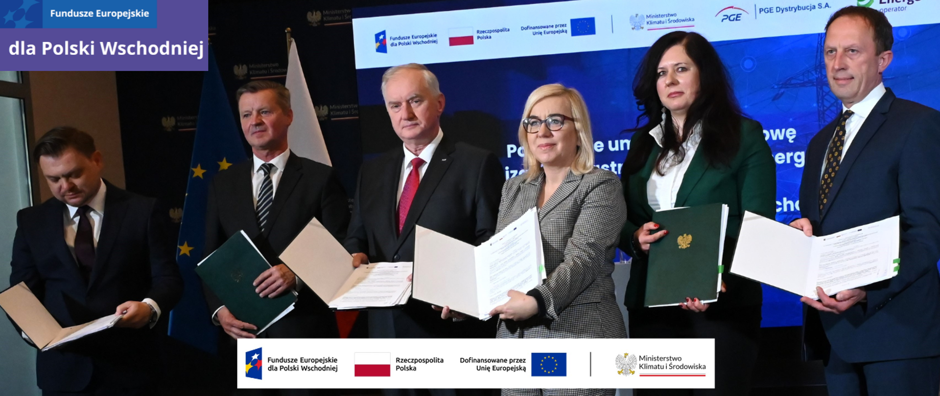Zdjęcie przedstawia sygnatariuszy umów na rozbudowę i modernizację infrastruktury elektroenergetycznej w ramach działania 2.1 Dystrybucja energii programu FEPW. W spotkaniu obok minister klimatu i środowiska Pauliny Hennig-Kloski uczestniczyli: Dariusz Marzec, prezes zarządu PGE SA, Jacek Drozd - prezes i Tomasz Małecki – wiceprezes zarządu PGE Dystrybucja, a także Marek Kasicki – wiceprezes zarządu i Karolina Błaszczak-Lasota – prokurent z Energa-Operator. Każdy z nich pokazuje podpisaną umowę. W tle jest telebim na którym widać granatową planszę z białym napisem: Podpisanie umów na rozbudowę i modernizację infrastruktury elektroenergetycznej w ramach działania 2.1 Fundusze Europejskie dla Polski Wschodniej. W lewym górnym rogu jest logo FEPW, na dole ciąg logotypów: znak FEPW, RP, UE oraz MKiŚ. 