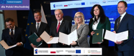 
Zdjęcie przedstawia sygnatariuszy umów na rozbudowę i modernizację infrastruktury elektroenergetycznej w ramach działania 2.1 Dystrybucja energii programu FEPW. Na zdjęciu stoją obok siebie: minister klimatu i środowiska Paulina Hennig-Kloska, Dariusz Marzec - prezes zarządu PGE SA, ze spółki PGE Dystrybucja: Jacek Drozd - prezes i Tomasz Małecki – wiceprezes zarządu, a ze spółki Energa-Operator: Marek Kasicki – wiceprezes zarządu i Karolina Błaszczak-Lasota – prokurent. Każdy z nich pokazuje podpisaną umowę. W tle jest telebim na którym widać granatową planszę z białym napisem: Podpisanie umów na rozbudowę i modernizację infrastruktury elektroenergetycznej w ramach działania 2.1 Fundusze Europejskie dla Polski Wschodniej. W lewym górnym rogu zdjęcia jest logo FEPW, na dole ciąg logotypów: znak FEPW, RP, UE oraz MKiŚ. 