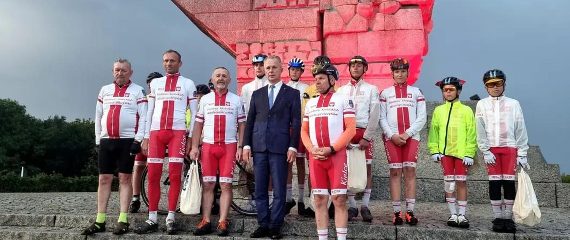 Grupa dzieci i dorosłych ubrana w kolarskie stroje pozuje na schodach przed pomnikiem na Westerplatte. Po środku mężczyzna w garniturze. 
