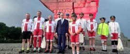 Grupa dzieci i dorosłych ubrana w kolarskie stroje pozuje na schodach przed pomnikiem na Westerplatte. Po środku mężczyzna w garniturze. 