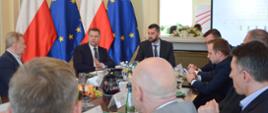 Uczestnicy spotkania siedzą i rozmawiają przy stole, za nimi flagi Polski i UE oraz duży ekran.