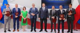 Zdjęcie zbiorowe uczestników uroczystości, stoją w rzędzie, za nimi wzór polskiej flagi i ekran z napisem Wręczenie powołań dla członków KPN.