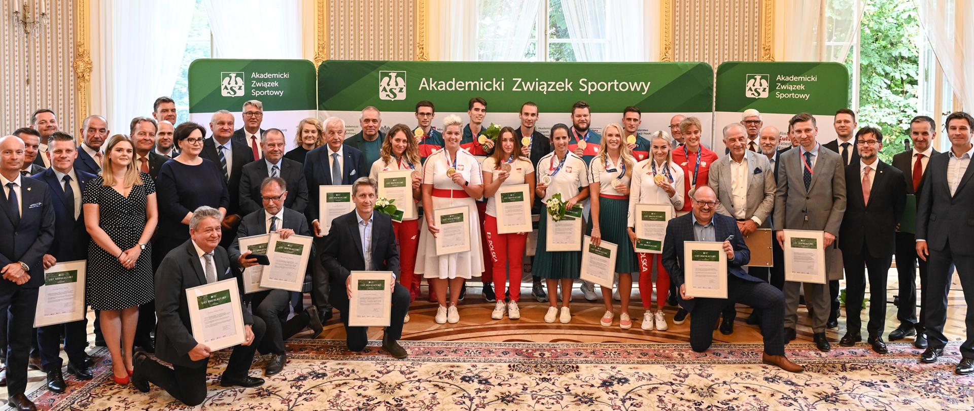 Na zdjęciu widoczni medaliści, trzymają w ręku medale oraz listy gratulacyjne na tle ścianki z napisem Akademicki Związek Sportowy, wokół medalistów stoją zebrani goście