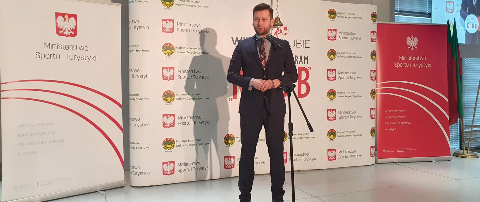 Rekrutacja do Rządowego Programu Klub rozpoczęta! Minister Kamil Bortniczuk przemawia do zgromadzonych gości, ogłaszając nabór wniosków do programu Klub. W tle ścianka Ministerstwa Sportu i Turystyki oraz Krajowego Zrzeszenia Ludowe Zespoły Sportowe