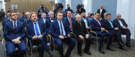 Widok od przodu na salę wypełnioną siedzącymi na ustawionych w rzędy krzesłach ludźmi w garniturach, za nimi kamery i mikrofony na wysięgnikach.
