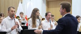 Kilkanaście młodych osób w białych koszulach gra na instrumentach dętych, przed nimi dyrygent w granatowym garniturze.