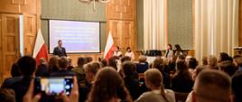 Widok od tyłu na wypełnioną siedzącymi na krzesłach ludźmi salę z wyłożonymi drewnianymi panelami ścianami, z przodu pod ścianą z wielkim ekranem stoi mężczyzna w garniturze i mówi do mikrofonu.