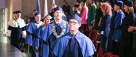 Ludzie w niebieskich ceremonialnych strojach idą rzędem przez salę, z boku widać krzesła.