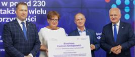 Na tle dużego ekranu stoi trzech mężczyzn i kobieta. Kobieta z mężczyzną trzyma dużą plansze z napisem Branżowe Centrum Umiejętności 10 mln zł. 