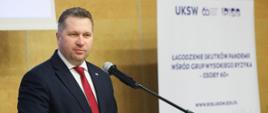 Minister Czarnek stoi przy mównicy i mówi do mikrofonu, za nim żółta ściana a przed nią baner z napisem UKSW - Łagodzenie skutków pandemii wśród grup wysokiego ryzyka.