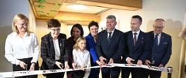 W rzędzie stoi grupa osób, cztery kobiety, trzech mężczyzn w garniturach i dziecko, wszyscy trzymają nożyki i przecinają wstęgę z napisami SOWA.