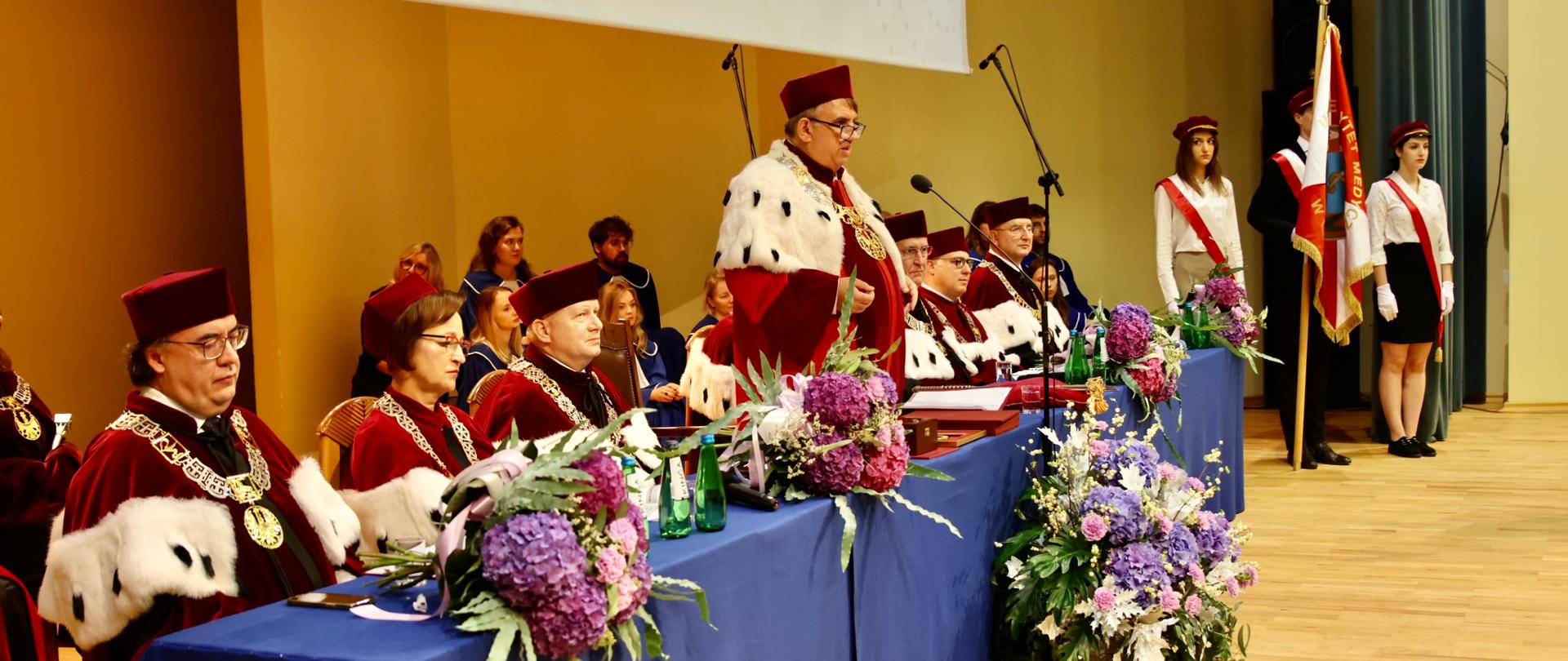 Widok z boku pod kątem na nakryty niebieskim materiałem stół za którym siedzą ludzie w czerwonych ceremonialnych strojach z gronostajowymi kołnierzami, na stole leżą bukiety kolorowych kwiatów, pośrodku stołu mężczyzna stoi i mówi do mikrofonu.