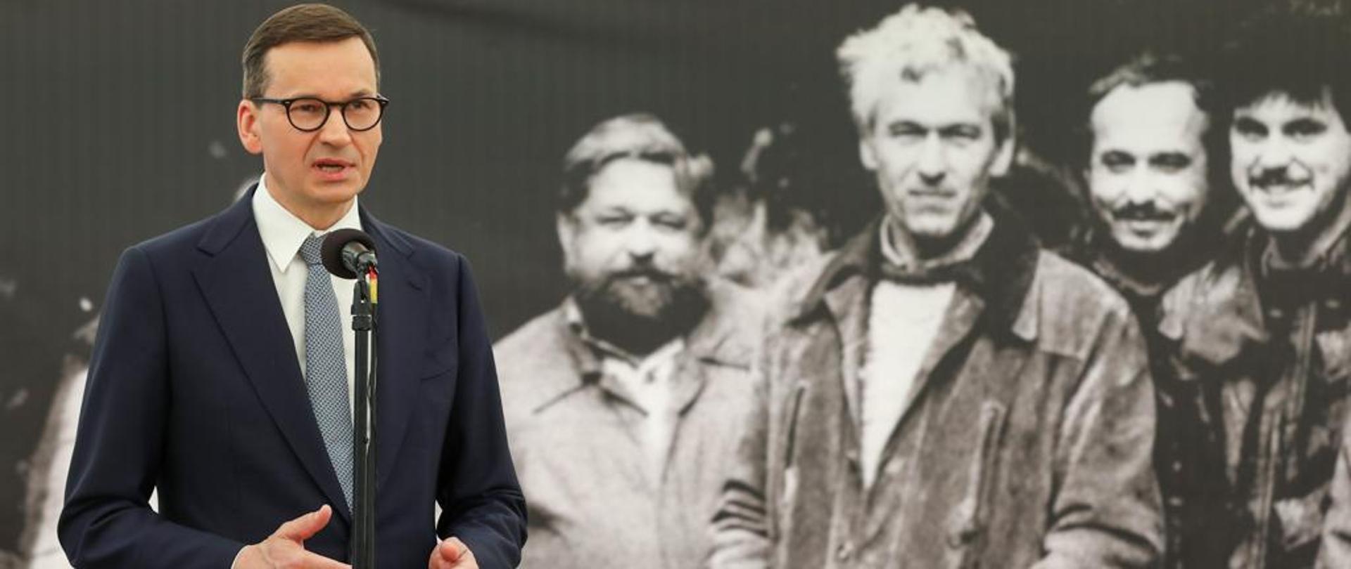 Premier Mateusz Morawiecki podczas uroczystości z okazji 40-lecia Solidarności Walczącej w Muzeum Żołnierzy Wyklętych i Więźniów Politycznych PRL. Premier stoi na scenie przed mikrofonem. W tle zdjęcie śp. Kornela Morawieckiego.