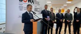 Pod dużym banerem z napisem SOWA stoi mężczyzna w garniturze i mówi do mikrofonu na stojaku, za nim pięciu mężczyzn w garniturach, dalej widać oświetlony korytarz.