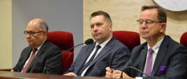 Za drewnianym blatem siedzi minister Czarnek i dwóch mężczyzn w garniturach, za nimi na żółtej ścianie biały herb przedstawiający dwa stojące lwy.
