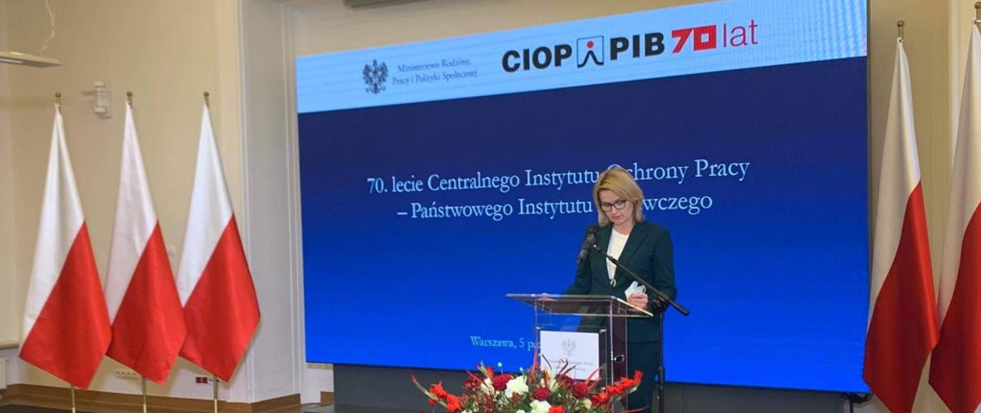 Sala. Minister Barbara Socha na uroczystości obchodów 70.lecia CIOP. Przemawia do zgromadzonych stojąc przy mównicy. Przed mównicą bukiet kolorowych kwiatów.