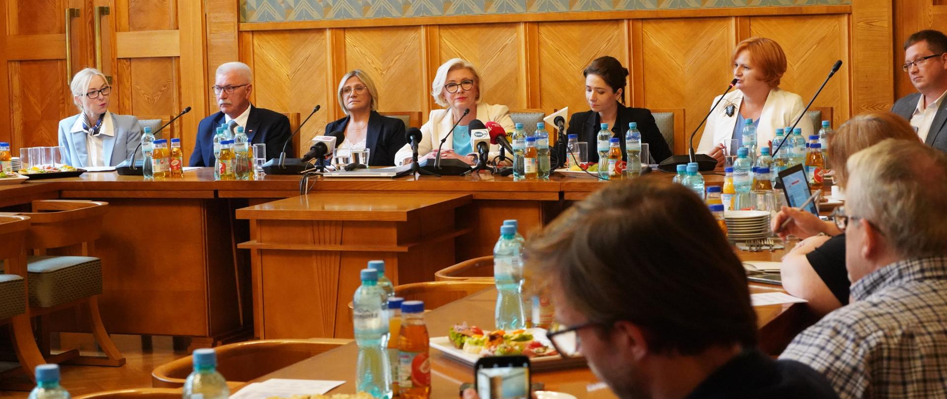 W sali ze ścianami wyłożonymi drewnianymi panelami przy ustawionych w czworokąt stołach siedzi dużo ludzi.