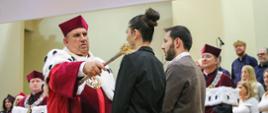 Mężczyzna w ceremonialnym czerwonym stroju trzyma w ręku drewniany kij, którym dotyka ramienia młodej stojącej przed nim kobiety, za nimi stoją ludzie.