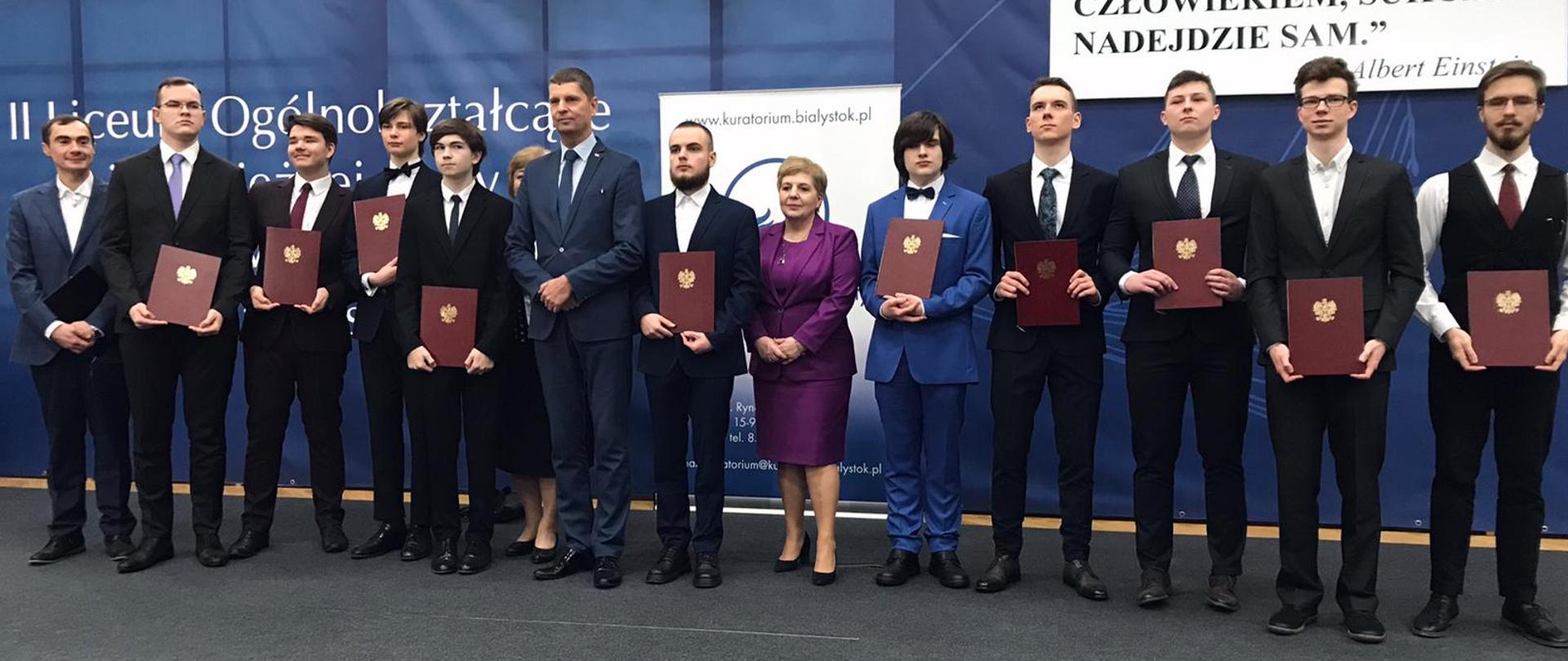 Wiceminister Piontkowski, kobieta ubrana na fioletowo i kilkunastu młodych ludzi w garniturach trzymających dyplomy w brązowych okładkach stoją w rzędzie pod granatową ścianą.