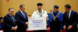Pod żółtą ścianą stoi pięciu mężczyzn, pośrodku grupy mężczyzna w ceremonialnym stroju z gronostajowym kołnierzem trzyma wielki symboliczny czek z napisem 25 500 000 zł.
