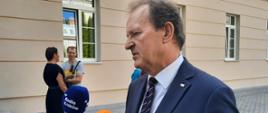 Zdjęcie z boku - na dworze przed budynkiem stoi wiceminister Bernacki który mówi do kilku mikrofonów trzymanych przez reporterów.