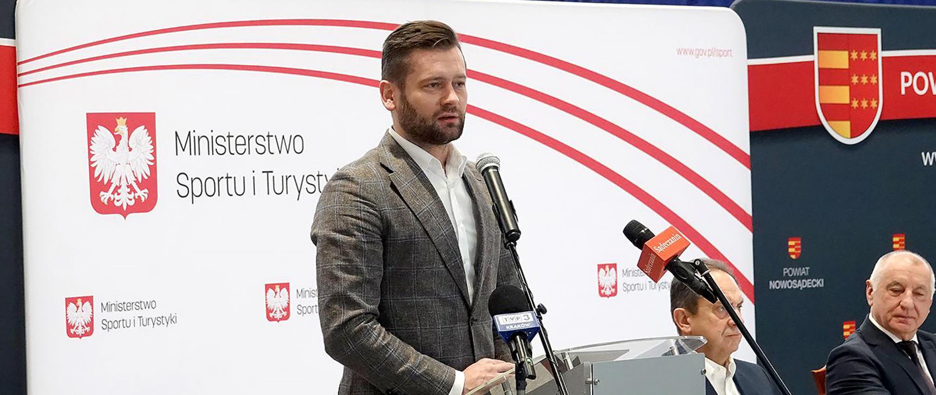 Spotkanie ministrów Bortniczuka i Guta-Mostowego z samorządowcami powiatu nowosądeckiego; minister Kamil Bortniczuk stoi przy mównicy, w tle ścianka z logotypami MSiT powiatu nowosądeckiego