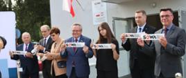 Przed wejściem do budynku stoi w rzędzie siedem osób, każdy trzyma kawałek wstęgi z napisem SOWA.